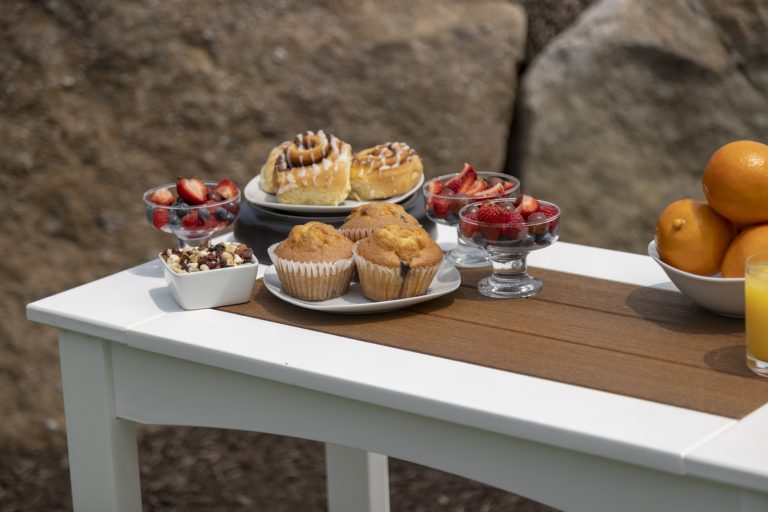 A table set outdoors features muffins, pastries, fruit cups, and oranges, with a glass of juice nearby.