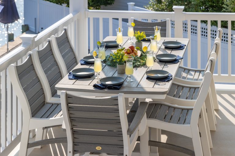 A beautifully set outdoor dining table with drinks, plates, and a centerpiece, surrounded by white chairs on a sunny balcony.