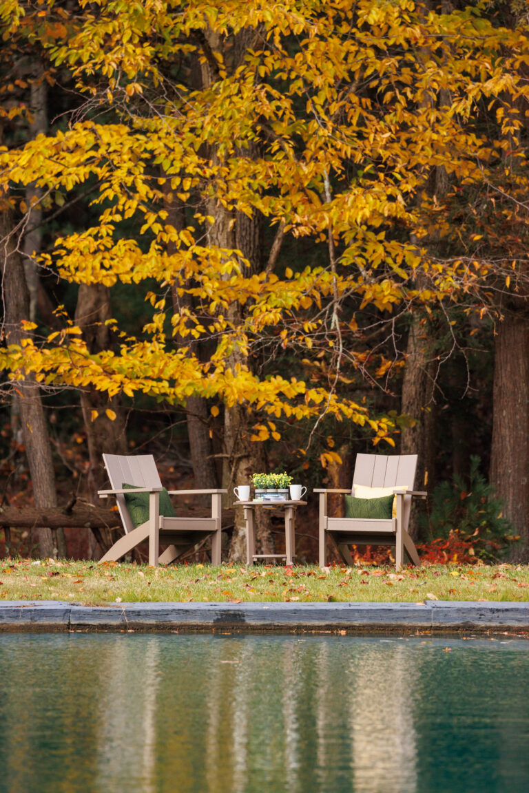 Two wooden chairs and a small table by a tranquil pond, surrounded by autumn foliage.