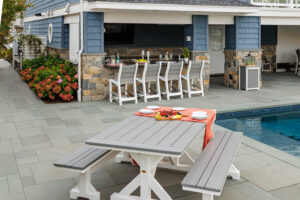 A patio scene featuring a white picnic table with a red tablecloth, surrounded by chairs and a pool in the background.