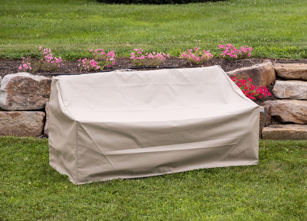 A beige outdoor furniture cover drapes over a couch, set on green grass near a stone wall and blooming pink flowers.